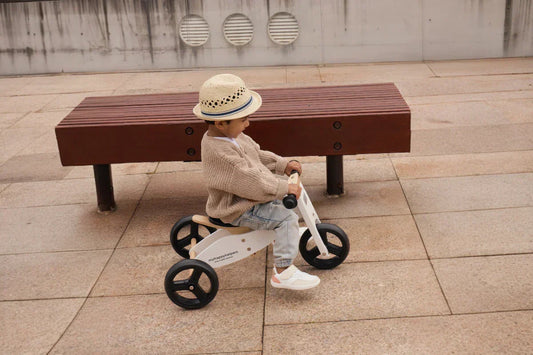Toddler in a straw hat rides a small white trike on a paved plaza, wearing a beige knit sweater, light jeans, and Rafferty’s Shoes in Desert Sand.