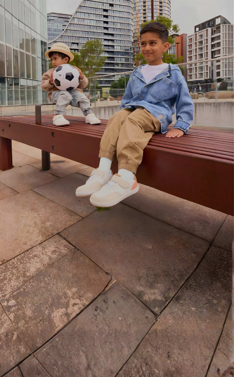 Two children sitting on a bench in a city plaza, wearing Rafferty’s kids barefoot sneakers; one holds a soccer ball.